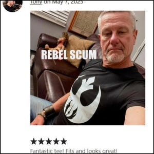 Pu&ograve; includere: T-shirt nera con grafica bianca e la scritta "REBEL SCUM". La grafica &egrave; un simbolo stilizzato. La persona nell'immagine indossa la maglietta.