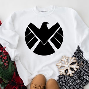 Puede incluir: Sudadera blanca con un gr&aacute;fico negro del logotipo de S.H.I.E.L.D. El logotipo es un c&iacute;rculo con un &aacute;guila negra en el interior y un escudo negro con tres l&iacute;neas horizontales.