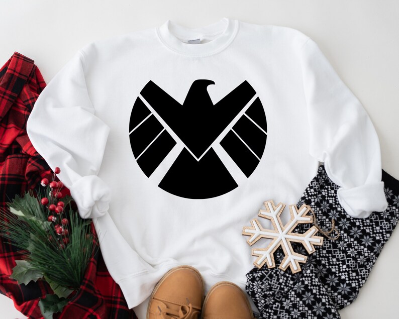 Puede incluir: Sudadera blanca con un gr&aacute;fico negro del logotipo de S.H.I.E.L.D. El logotipo es un c&iacute;rculo con un &aacute;guila negra en el interior y un escudo negro con tres l&iacute;neas horizontales.