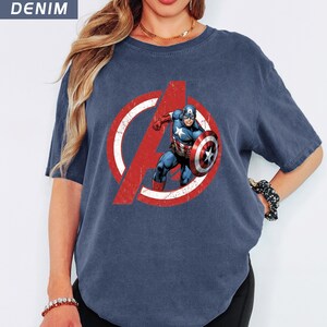 Puede incluir: Camiseta azul denim con un gr&aacute;fico del Capit&aacute;n Am&eacute;rica dentro de un logotipo rojo y blanco de los Vengadores. El superh&eacute;roe se muestra con su escudo. La camiseta tiene un aspecto vintage y descolorido. La palabra 'DENIM' es visible.