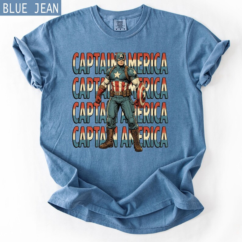 May include: A blue jean colored t-shirt featuring a graphic of Captain America. The design includes the words "CAPTAIN AMERICA" stacked vertically in a retro style, with the superhero in the center, holding his shield.