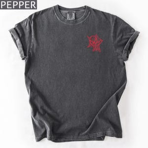 K&ouml;nnte beinhalten: Dunkelgraues T-Shirt mit roter Spinnennetz- und Spinnen-Grafik auf der Vorderseite. Es hat einen Rundhalsausschnitt und kurze &Auml;rmel. Das Wort "PEPPER" befindet sich in der oberen linken Ecke.