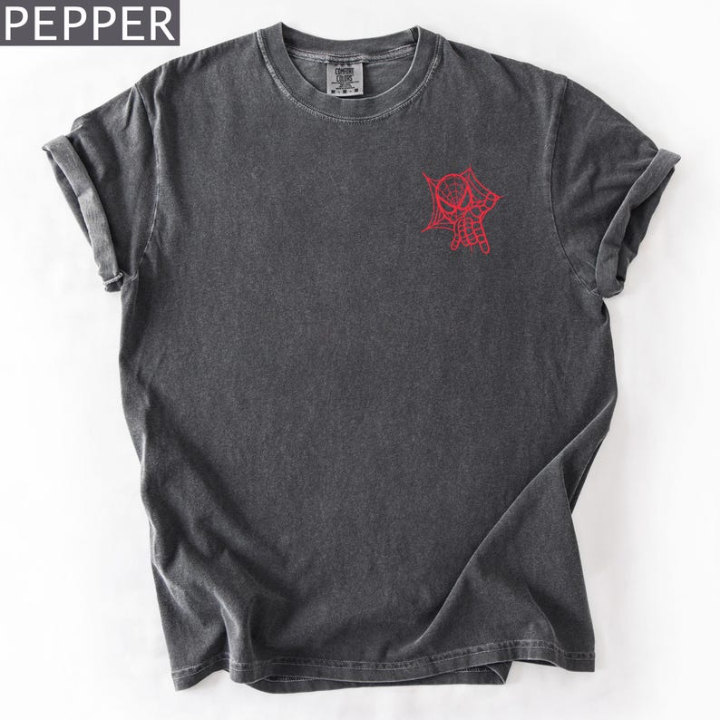 K&ouml;nnte beinhalten: Dunkelgraues T-Shirt mit roter Spinnennetz- und Spinnen-Grafik auf der Vorderseite. Es hat einen Rundhalsausschnitt und kurze &Auml;rmel. Das Wort "PEPPER" befindet sich in der oberen linken Ecke.