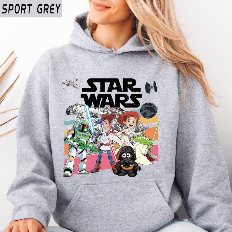 Puede incluir: Sudadera gris deportiva con un gr&aacute;fico colorido de Star Wars. El dise&ntilde;o presenta personajes como Darth Vader, Woody y Buzz Lightyear. El logotipo de Star Wars es visible. La sudadera tiene capucha con cord&oacute;n y bolsillo canguro.