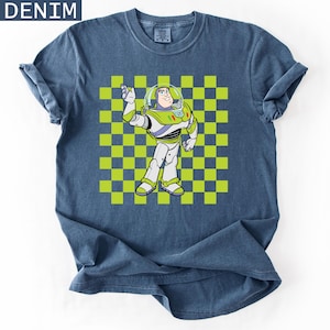 May include: Denim blue t-shirt featuring a graphic of Buzz Lightyear from Toy Story. The character is in a white, green, and purple spacesuit, set against a green and yellow checkered background. The word "DENIM" is in the top left corner.
