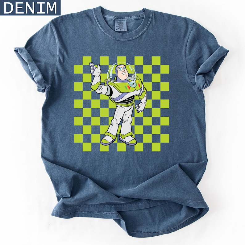 May include: Denim blue t-shirt featuring a graphic of Buzz Lightyear from Toy Story. The character is in a white, green, and purple spacesuit, set against a green and yellow checkered background. The word "DENIM" is in the top left corner.
