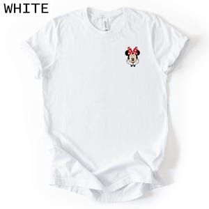 May include: A white short-sleeved t-shirt featuring a small graphic of Minnie Mouse on the upper left chest area. The character is depicted with her signature red and white polka dot bow, black ears, and white gloves resting near her face.