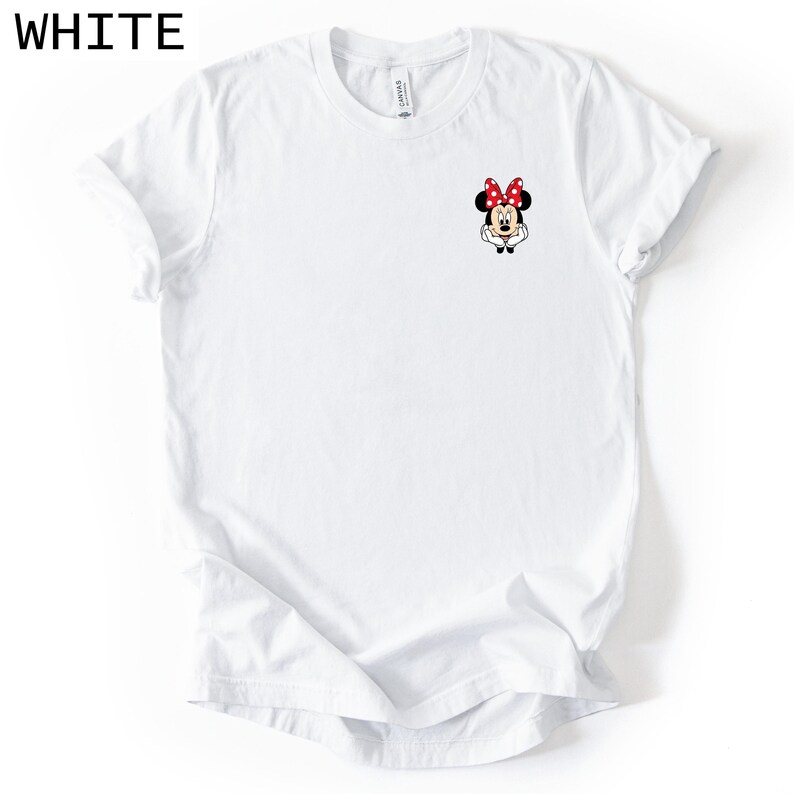 May include: A white short-sleeved t-shirt featuring a small graphic of Minnie Mouse on the upper left chest area. The character is depicted with her signature red and white polka dot bow, black ears, and white gloves resting near her face.