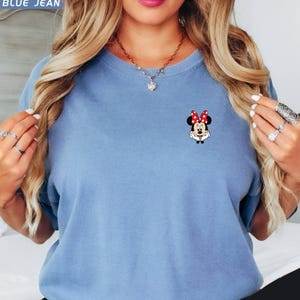 May include: A light blue t-shirt with a small Minnie Mouse graphic on the chest. The graphic shows Minnie Mouse with a red polka dot bow. The shirt has short sleeves and a crew neck.