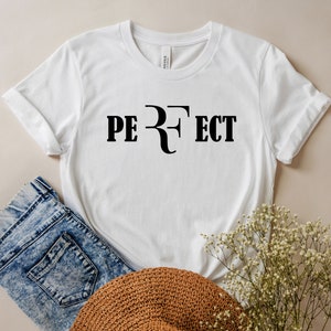 Op de afbeelding: Wit T-shirt met zwarte tekst die "PERFECT" luidt met een gestileerde "R" in het midden.