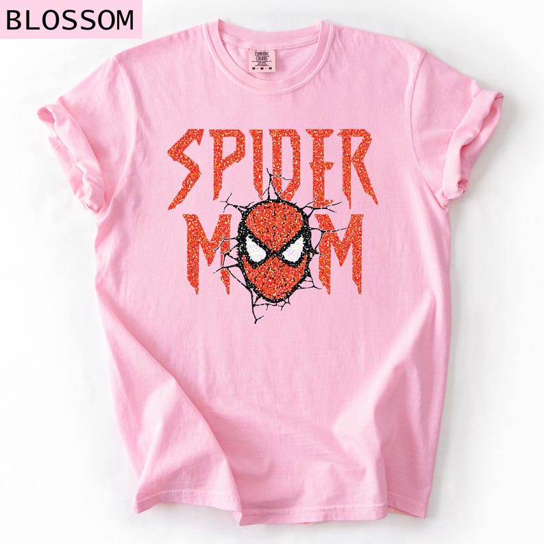 May include: A light pink t-shirt with the words "SPIDER MOM" in orange glitter lettering. The design includes a Spider-Man mask in the center, with a cracked effect around it. The word "BLOSSOM" is in the top left corner.