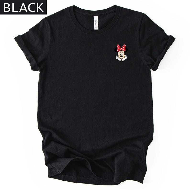 May include: A black short-sleeved t-shirt featuring a small graphic of Minnie Mouse on the upper left chest. The character is depicted with a red and white polka-dot bow, white gloves, and hands resting under the chin. The word BLACK is in the corner.