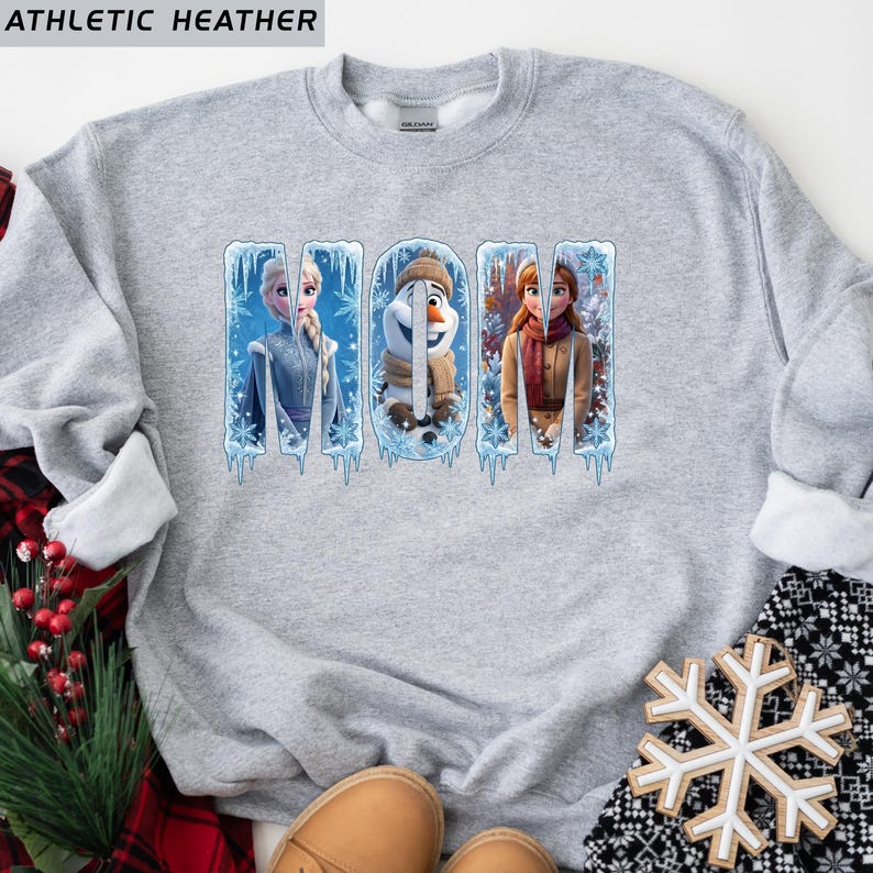 Puede incluir: Sudadera gris jaspeado con la palabra "MOM" en letras azul hielo. Las letras presentan im&aacute;genes de personajes de la pel&iacute;cula Frozen. La esquina superior izquierda de la sudadera tiene el texto "ATHLETIC HEATHER".