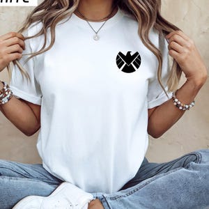 May include: White t-shirt with a black graphic of a stylized bird emblem on the chest. The shirt has short sleeves and a crew neck. The word "HITE" is in the top left corner.