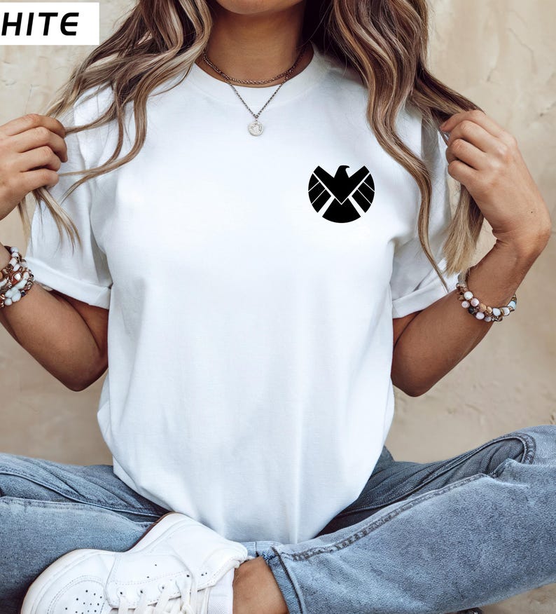 May include: White t-shirt with a black graphic of a stylized bird emblem on the chest. The shirt has short sleeves and a crew neck. The word "HITE" is in the top left corner.
