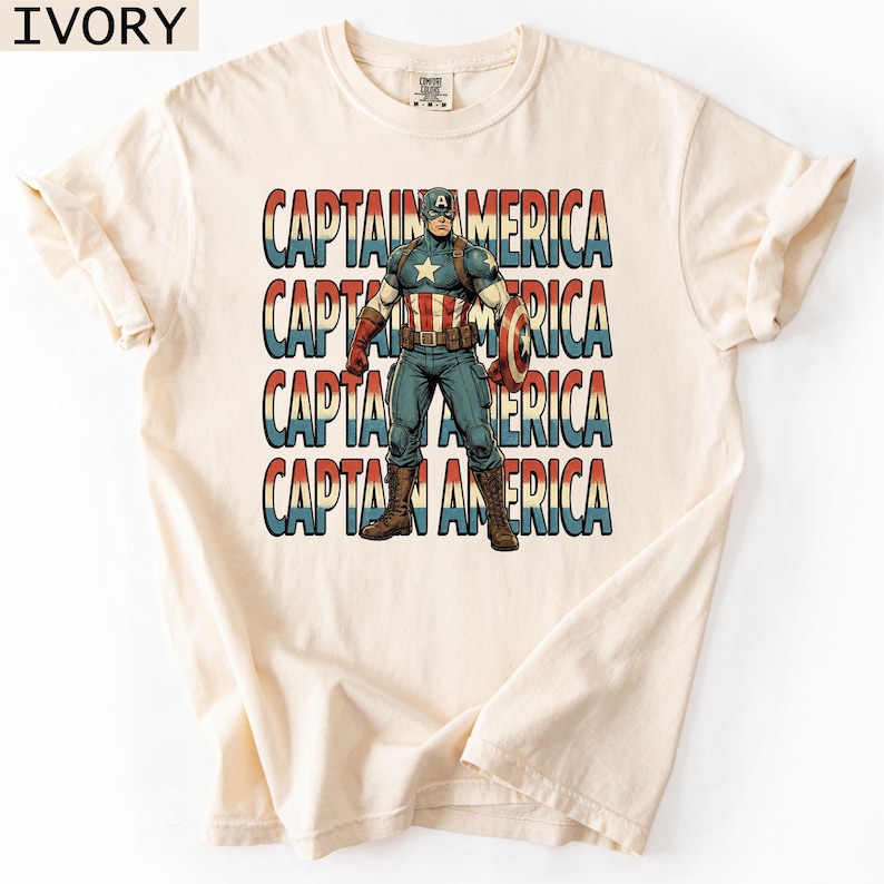 May include: Ivory-colored t-shirt featuring a graphic of Captain America in a vintage style. The design includes the text "CAPTAIN AMERICA" stacked four times in red, white, and blue. The superhero is depicted with his shield.