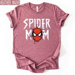 May include: Heather mauve t-shirt featuring the text "SPIDER MOM" in white, and a red and black spider mask graphic. The shirt has a crew neck and is made of a soft material. The text "HEATHER MAUVE" is in the top left corner.