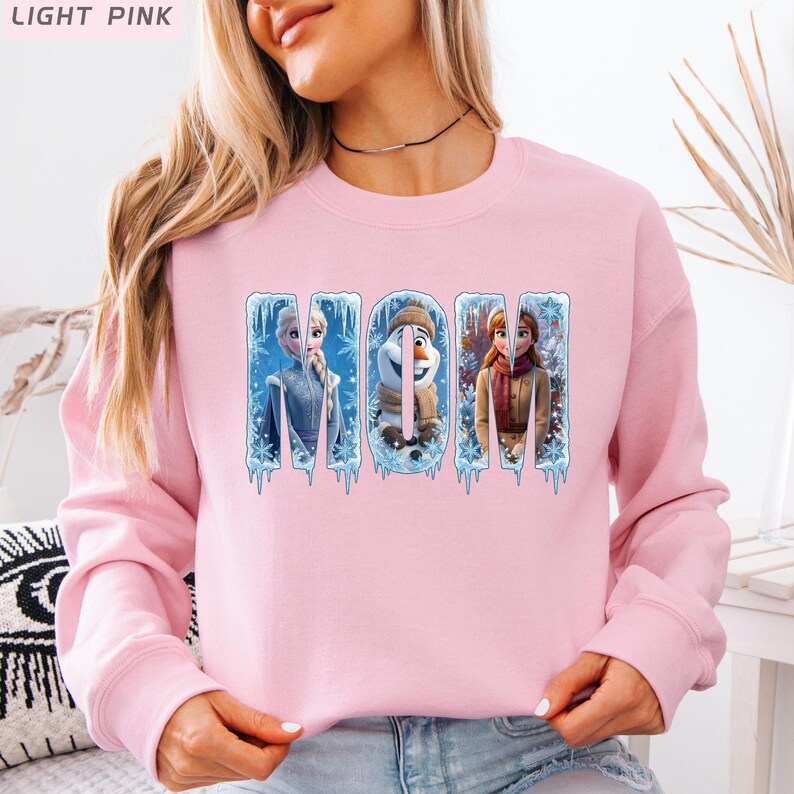 Puede incluir: Sudadera rosa claro con la palabra "MOM" en un dise&ntilde;o de tem&aacute;tica invernal. Las letras est&aacute;n decoradas con im&aacute;genes de personajes de la pel&iacute;cula Frozen, incluyendo Elsa, Olaf y Anna. La sudadera es de cuello redondo.