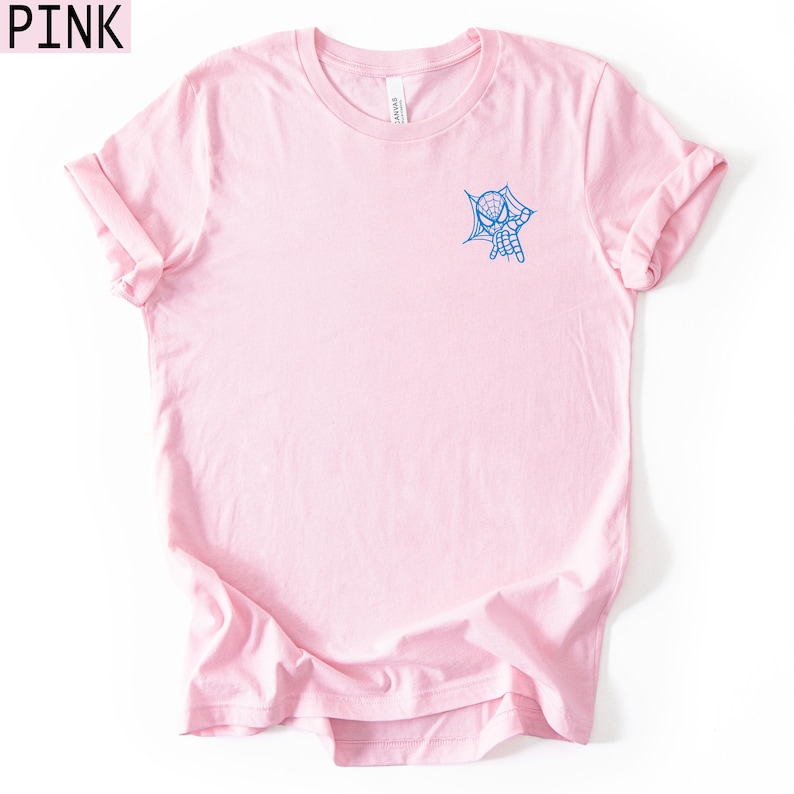 Op de afbeelding: Een lichtroze T-shirt met korte mouwen en een kleine blauwe afbeelding op de linkerborst. De afbeelding toont een masker in spinnenthema dat uit een webontwerp tevoorschijn komt. Het T-shirt heeft een ronde hals en een standaard pasvorm.