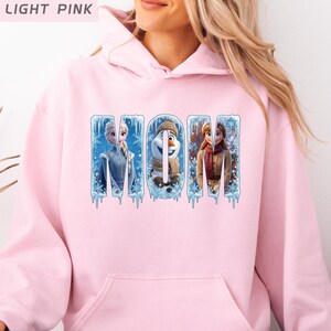 Puede incluir: Sudadera con capucha rosa claro con la palabra "MOM" en una fuente azul hielo. Las letras presentan im&aacute;genes de personajes de dibujos animados. La sudadera tiene un bolsillo delantero y una capucha.