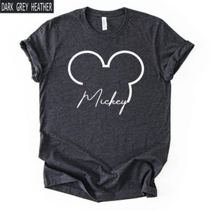 May include: Dark grey heather t-shirt with a white outline of Mickey Mouse ears and the name "Mickey" in a cursive font. The shirt has rolled-up sleeves and a crew neck.