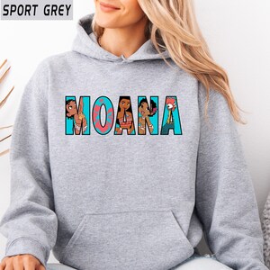 May include: Sport grey hoodie with the word "MOANA" in a colourful design. The letters are filled with cartoon characters. The hoodie has a front pocket and a drawstring hood.