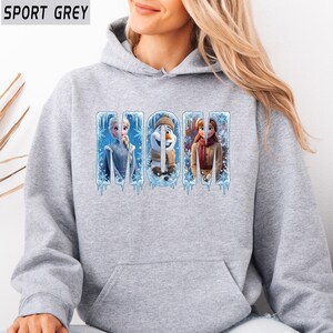 Puede incluir: Una sudadera con capucha gris claro con la palabra "MOM" en un dise&ntilde;o de hielo azul. Las letras presentan personajes de la pel&iacute;cula Frozen: Elsa, Olaf y Anna. La sudadera est&aacute; etiquetada como "SPORT GREY" en la esquina superior izquierda.