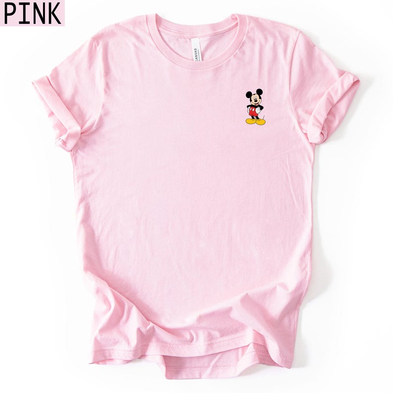 May include: A light pink short-sleeved t-shirt featuring a small graphic of Mickey Mouse on the upper left chest. The character is depicted with black ears, a red pair of shorts, and yellow shoes. The word PINK is printed in the top left corner.
