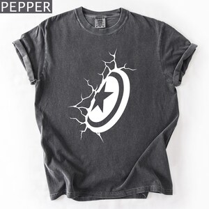 Pode incluir: Camiseta cinza escuro com um design branco de escudo e estrela. O gr&aacute;fico parece estar quebrando a camiseta, com linhas de rachaduras que emanam do escudo. A palavra "PEPPER" est&aacute; no canto superior esquerdo.