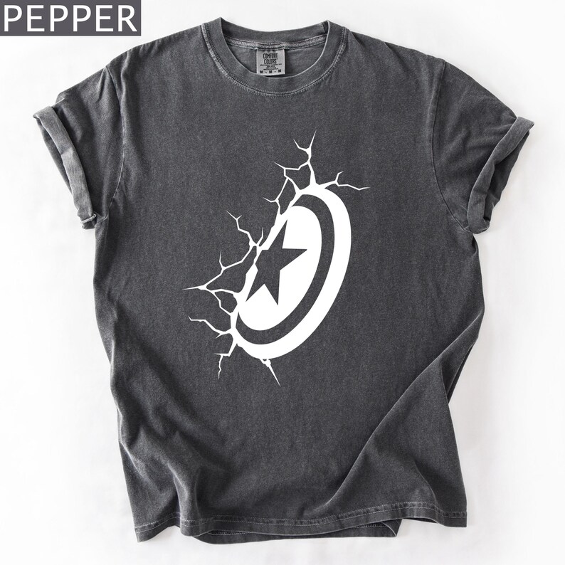 Pode incluir: Camiseta cinza escuro com um design branco de escudo e estrela. O gr&aacute;fico parece estar quebrando a camiseta, com linhas de rachaduras que emanam do escudo. A palavra "PEPPER" est&aacute; no canto superior esquerdo.