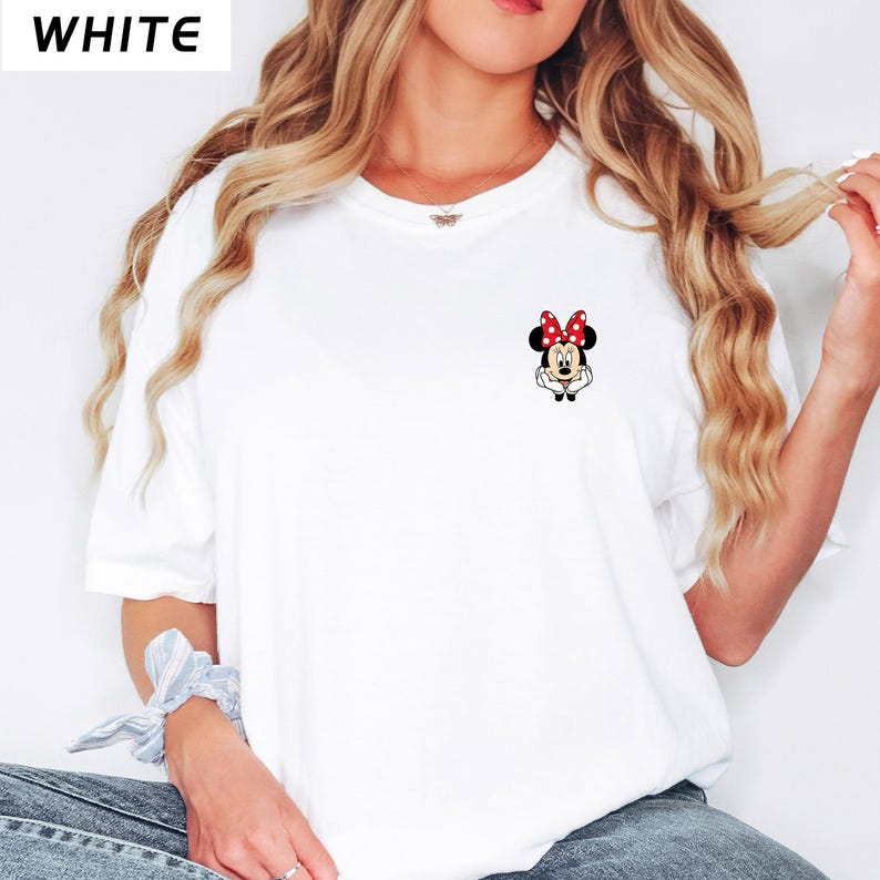May include: White t-shirt featuring a small Minnie Mouse graphic on the chest. The cartoon character wears a red bow with white polka dots. The word "WHITE" is printed in black in the upper left corner of the image.