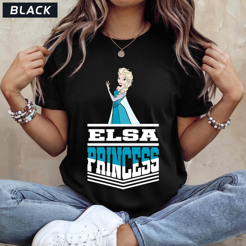 May include: Black t-shirt featuring a graphic of Elsa from Frozen, with the text "ELSA PRINCESS" in white and blue. The shirt has a crew neck and short sleeves. The word "BLACK" is in a white box in the upper left corner.