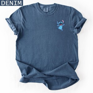 May include: Denim blue t-shirt featuring a small embroidered image of the character Stitch. The shirt has rolled-up sleeves and the word "DENIM" in a white box in the upper left corner. The shirt is made of a soft, comfortable material.