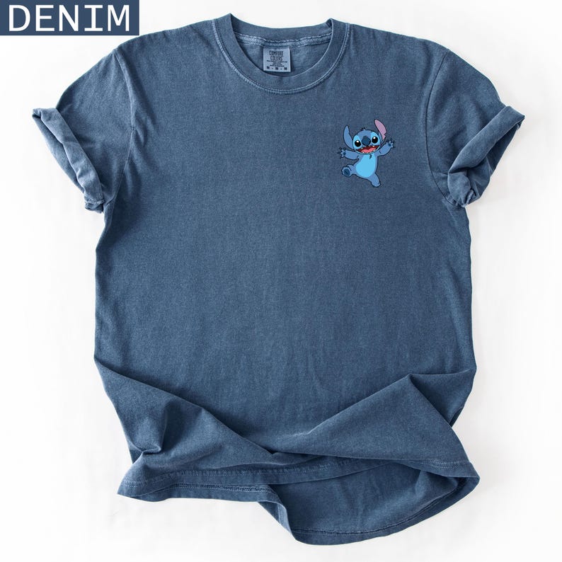 May include: Denim blue t-shirt featuring a small embroidered image of the character Stitch. The shirt has rolled-up sleeves and the word "DENIM" in a white box in the upper left corner. The shirt is made of a soft, comfortable material.