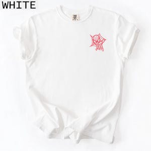 K&ouml;nnte beinhalten: Wei&szlig;es T-Shirt mit rotem Spider-Man-Netz- und Handdesign auf der Vorderseite. Das Wort "WHITE" befindet sich in der oberen linken Ecke. Das Shirt hat einen Rundhalsausschnitt und kurze &Auml;rmel.