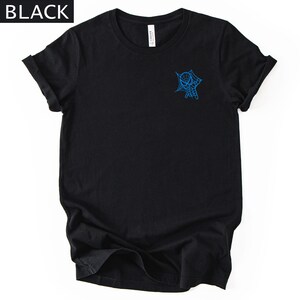 Op de afbeelding: Een zwart T-shirt met korte mouwen en een kleine blauwe afbeelding op de linkerborst. De afbeelding toont een karaktermasker dat uit een spinnenwebontwerp tevoorschijn komt. Het woord BLACK staat in de linkerbovenhoek van de afbeelding.