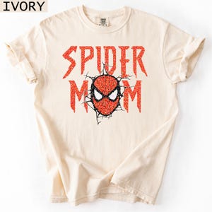 May include: Ivory-colored t-shirt with the words "SPIDER MOM" in red glitter lettering. The design includes a red and black spider mask graphic. The shirt is a comfortable, casual style.