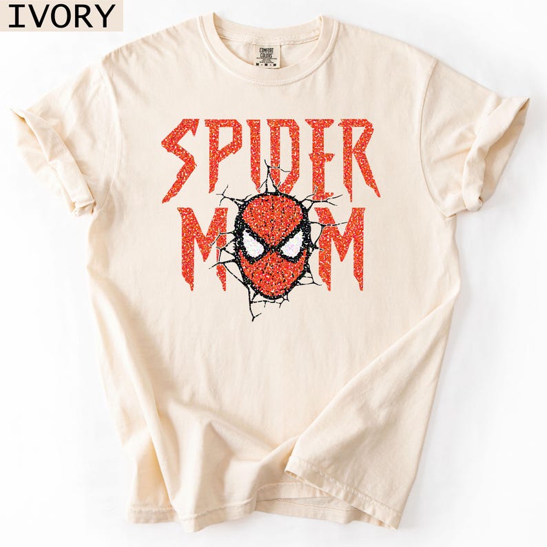 May include: Ivory-colored t-shirt with the words "SPIDER MOM" in red glitter lettering. The design includes a red and black spider mask graphic. The shirt is a comfortable, casual style.