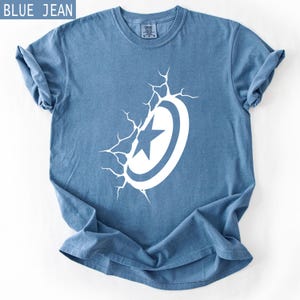 Pode incluir: T-shirt azul jeans com um desenho branco de escudo e estrela. O gr&aacute;fico do escudo parece estar a romper a superf&iacute;cie da camisa. A camisa tem mangas enroladas e gola redonda.