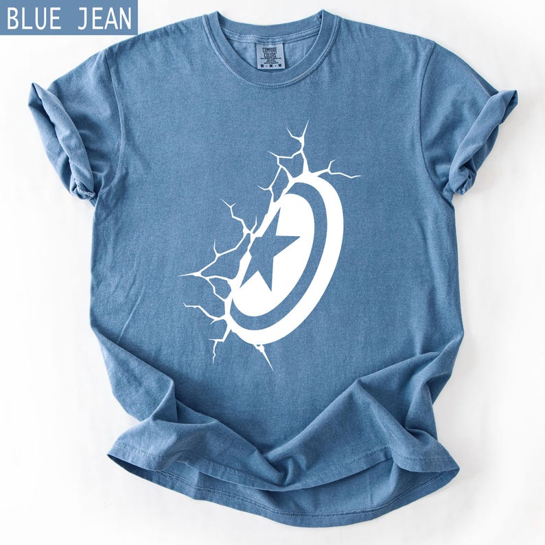 Pode incluir: T-shirt azul jeans com um desenho branco de escudo e estrela. O gr&aacute;fico do escudo parece estar a romper a superf&iacute;cie da camisa. A camisa tem mangas enroladas e gola redonda.