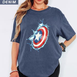 May include: A denim blue t-shirt featuring a graphic of Captain America's shield. The shield is red, white, and blue, with a white star in the center. The shield appears to be breaking through a splash of water.