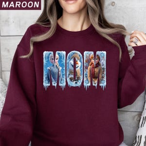 Puede incluir: Sudadera granate con la palabra "MOM" en un dise&ntilde;o de invierno. Las letras presentan im&aacute;genes de los personajes de la pel&iacute;cula Frozen: Elsa, Olaf y Anna. La sudadera es de color granate liso.