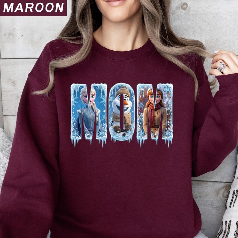Puede incluir: Sudadera granate con la palabra "MOM" en un dise&ntilde;o de invierno. Las letras presentan im&aacute;genes de los personajes de la pel&iacute;cula Frozen: Elsa, Olaf y Anna. La sudadera es de color granate liso.