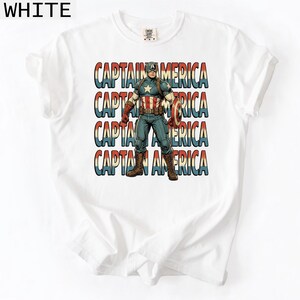May include: White t-shirt featuring a Captain America graphic. The design includes the text "CAPTAIN AMERICA" stacked vertically in red, white, and blue. The illustration depicts Captain America in his iconic costume, holding his shield.