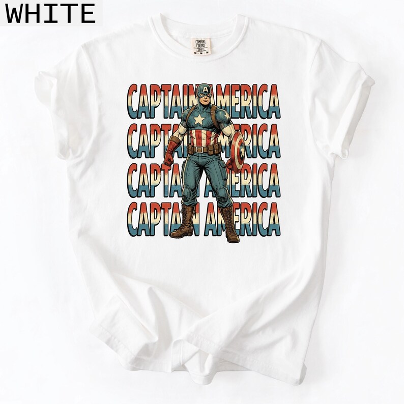 May include: White t-shirt featuring a Captain America graphic. The design includes the text "CAPTAIN AMERICA" stacked vertically in red, white, and blue. The illustration depicts Captain America in his iconic costume, holding his shield.