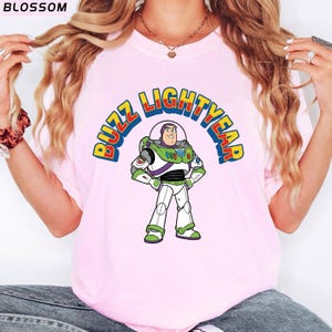 Op de afbeelding: Lichtroze T-shirt met een cartoonafbeelding van Buzz Lightyear uit Toy Story. De afbeelding toont de woorden "BUZZ LIGHTYEAR" in een kleurrijke boog boven het personage. Het personage draagt zijn groene, witte en paarse ruimtepak.