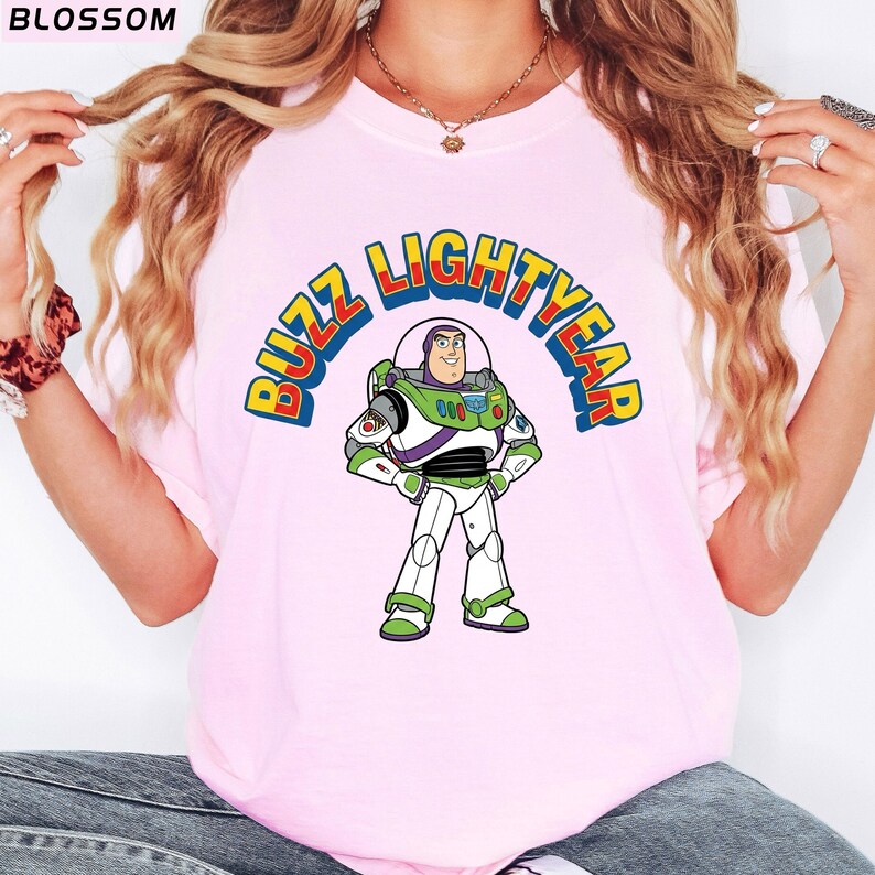 Op de afbeelding: Lichtroze T-shirt met een cartoonafbeelding van Buzz Lightyear uit Toy Story. De afbeelding toont de woorden "BUZZ LIGHTYEAR" in een kleurrijke boog boven het personage. Het personage draagt zijn groene, witte en paarse ruimtepak.