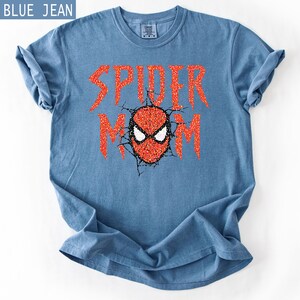 May include: Blue jean colored t-shirt with the words "SPIDER MOM" in red glitter lettering. The design includes a red and black spider mask graphic. The shirt is folded on a white surface.