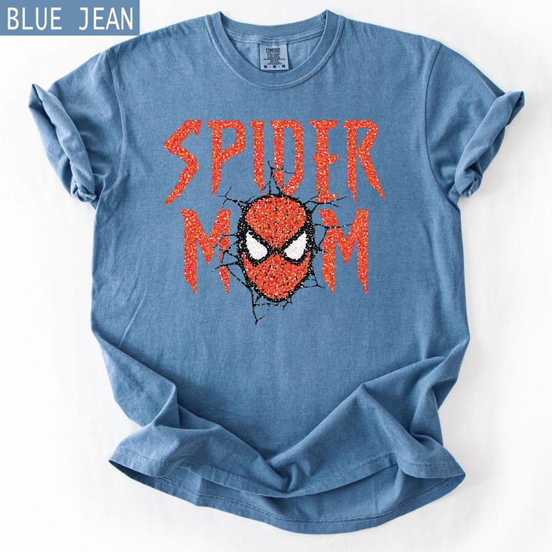 May include: Blue jean colored t-shirt with the words "SPIDER MOM" in red glitter lettering. The design includes a red and black spider mask graphic. The shirt is folded on a white surface.