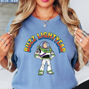 Op de afbeelding: Blauw spijkerbroekkleurig T-shirt met een afbeelding van Buzz Lightyear uit Toy Story. Het ontwerp bevat de afbeelding van het personage en de woorden "BUZZ LIGHTYEAR" in een kleurrijk, gebogen lettertype. Het shirt is effen van kleur en heeft korte mouwen.